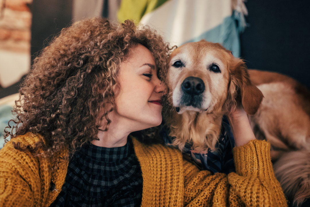 Woman with curly hair wearing a mustard cardigan kissing a golden retriever.